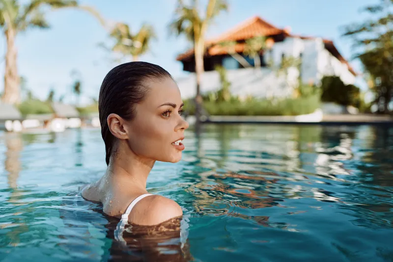 Young woman in white swimsuit relaxing in outdoor pool, calm water surface, tropical background, natural light, serene expression, summer lifestyle, sunny day