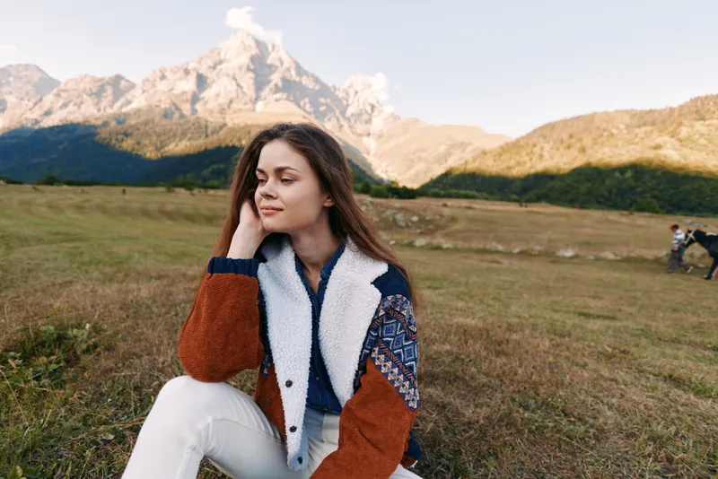 Woman portrait in mountains meadow, serene young traveler sitting on grass wearing warm jacket, enjoying nature and autumn scenery with distant peaks and soft sunlight.
