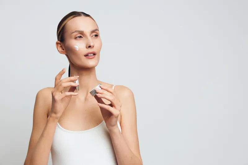 Young woman with clear skin applying white face cream on cheeks holding cosmetic jar, isolated on white background. Concept of skincare, beauty routine, and self-care with soft lighting.