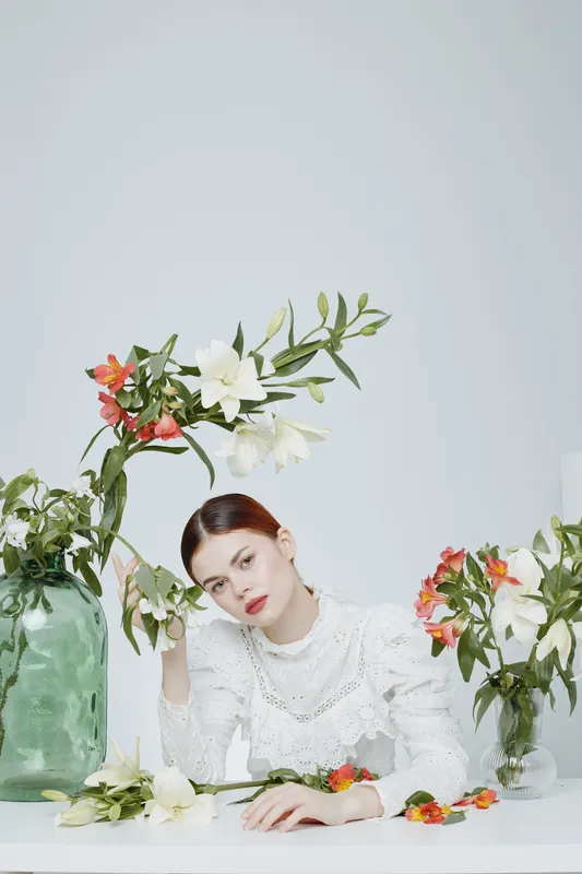 A serene young woman in a white blouse surrounded by blooming flowers in soft pastel hues, showcasing beauty and tranquility in an elegant setting