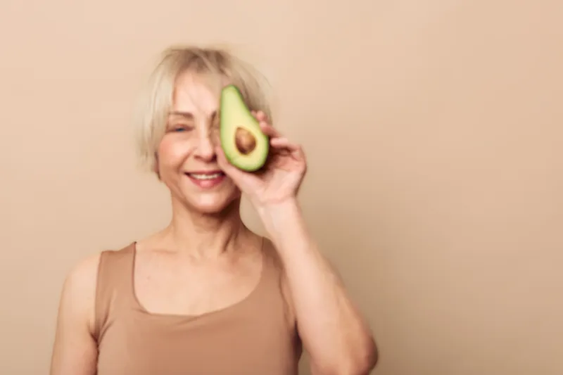 Smiling middle aged woman holds fresh avocado half close to face against beige background. Concept of healthy lifestyle, nutrition, natural beauty, wellness, and organic food.