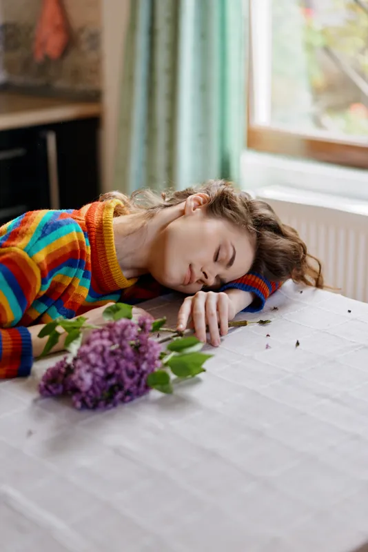Sleeping woman resting at a table indoors with a bouquet nearby, presenting a calm lifestyle moment, cozy wellbeing, and gentle relaxation under soft natural light.