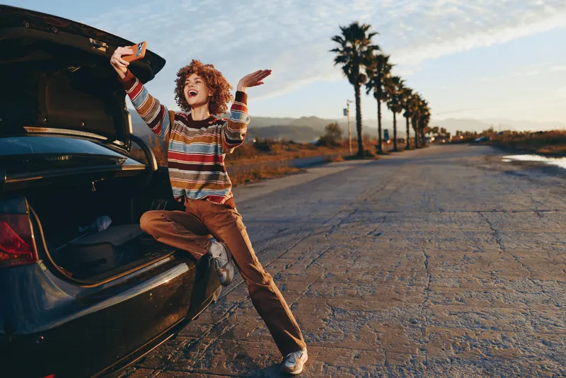 Woman lifestyle, near open car trunk on a road at sunset, holding a smartphone, laughing with joy, relaxed posture, warm outdoor travel light, cinematic everyday free moment