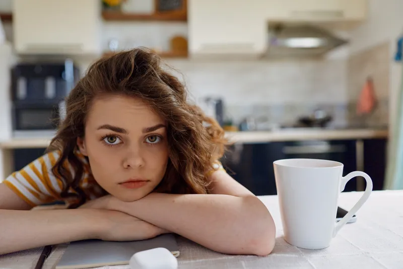 A young woman with wavy hair rests her head on a kitchen table, eyes tired, beside a white mug and laptop, conveying intimate quiet home moments and contemplation.