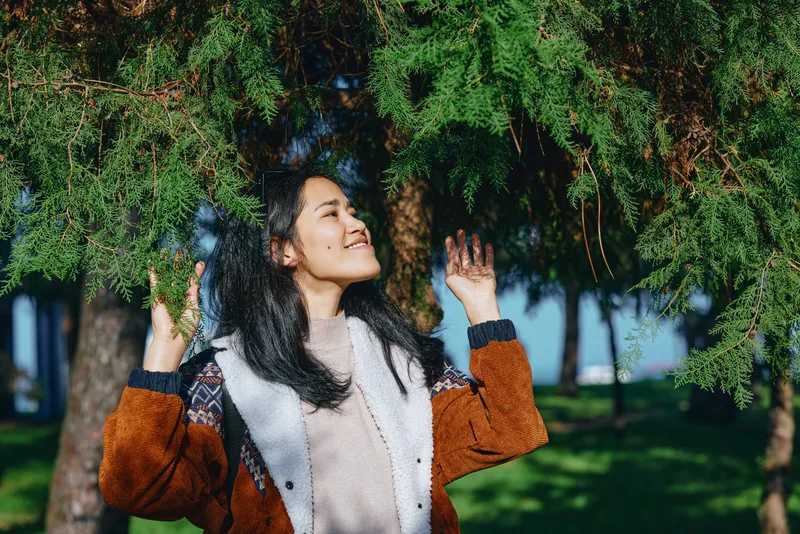 Outdoor scene with a smiling woman beneath hanging branches, arms raised, enjoying sunlight in a lush park, conveying happiness and nature connection for lifestyle and wellness themes