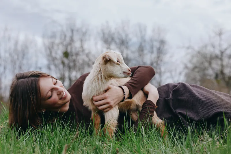 Cheerful woman outdoors lying on grass, embracing a young goat kid, warm sunlight, relaxed mood, rustic setting, soft breeze, close connection with nature, playful, spontaneous moment, genuine bond