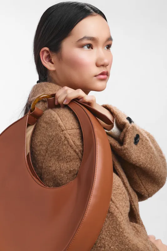 asian woman is photographed in a minimalist studio, calmly carrying a large leather bag over her shoulder. her poised pose and soft expression create a chic, contemporary fashion moment.