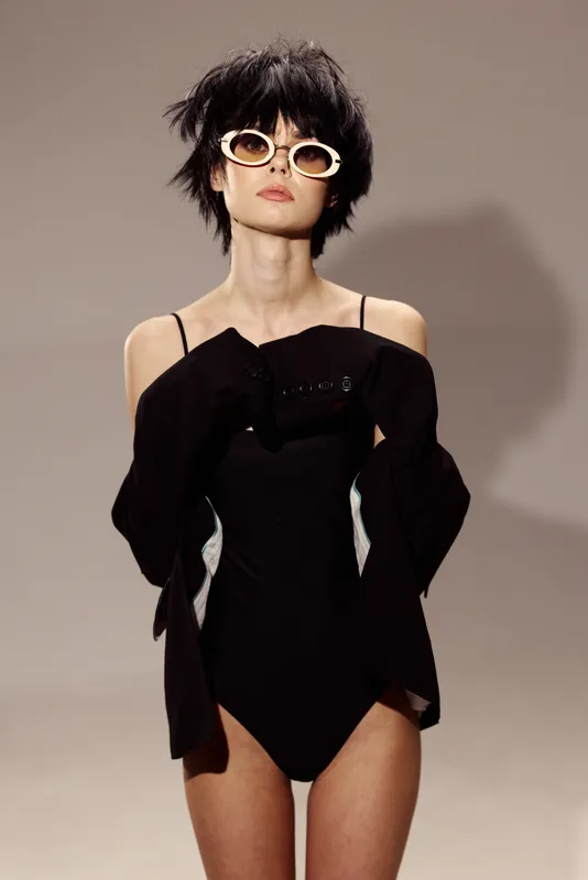 Woman in black bodysuit and white sunglasses posing with fashionable attitude on beige background.