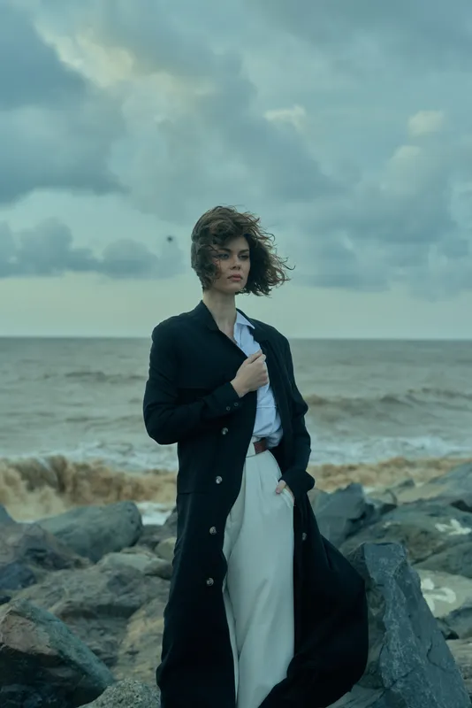 Woman in a long dark coat and white trousers stands on a rocky shoreline, wind-swept hair, posing with the coat open, moody ocean backdrop, fashion mood, cinematic style, outdoor shoot with dramatic