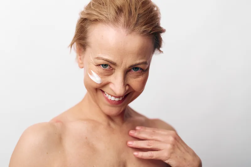 Smiling middle aged woman with natural skin applying white facial cream on cheek. Close up on white background. Concept of skincare, beauty, self care, and healthy aging.