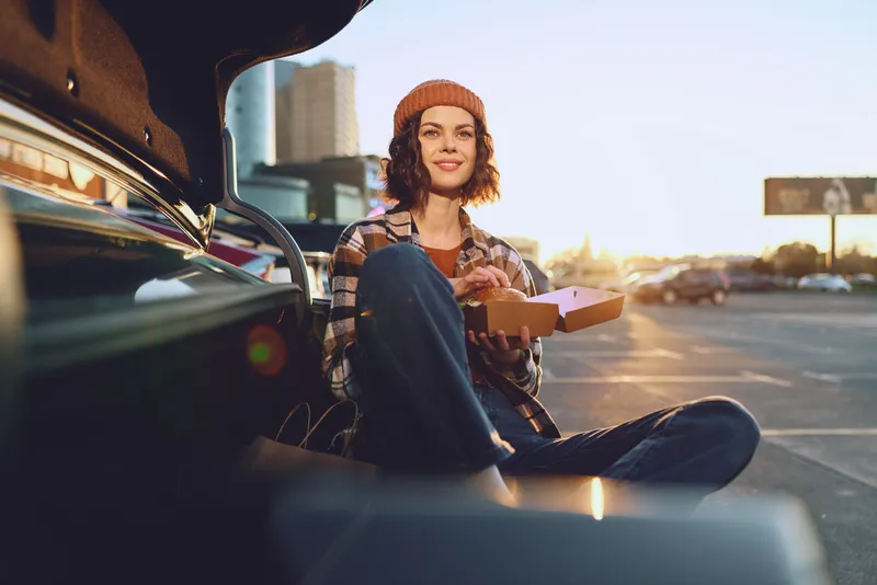Woman sitting at car trunk in parking at sunset eating takeaway food with a warm smile and beanie, candid lifestyle scene with authenticity, golden hour glow and mindful living.