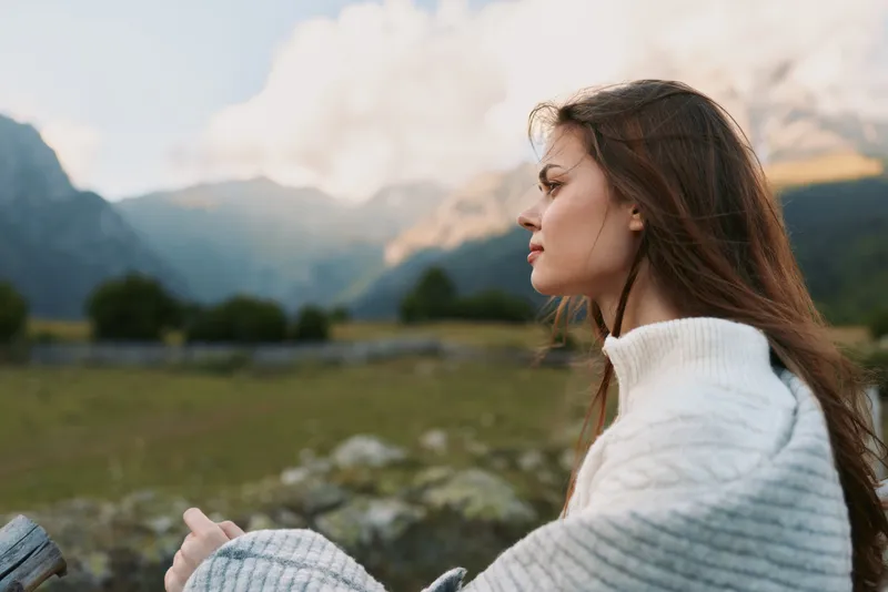 Young woman wrapped in blanket sits outdoors in mountain landscape looks thoughtfully into distance enjoys peaceful nature moment lifestyle concept.