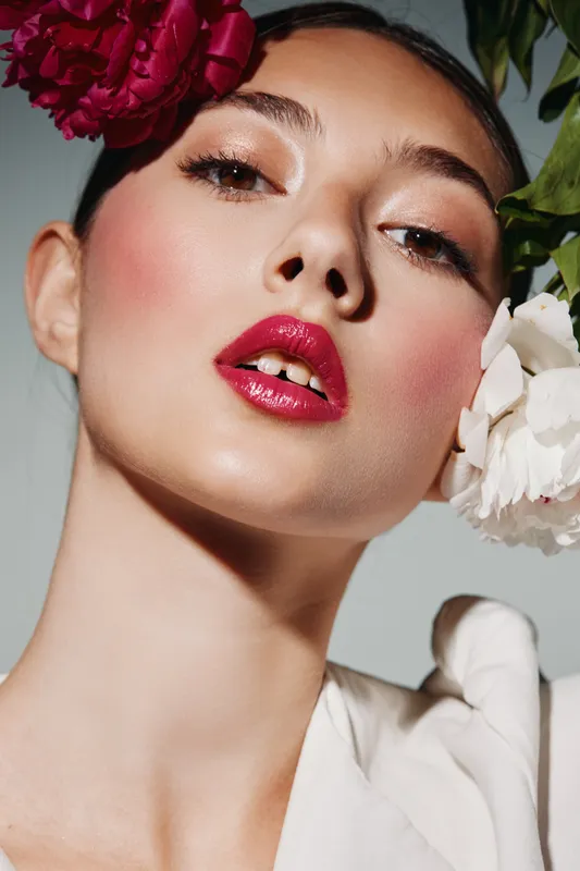 Woman with creative makeup and glossy red lips posing with bold floral accessories accentuating fresh flawless skin and modern fashion style on neutral backdrop.