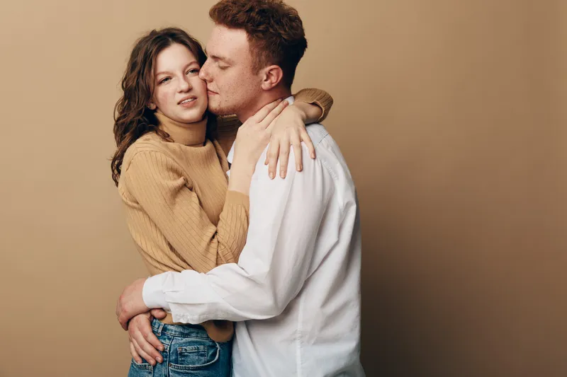 Young couple embraces tenderly against beige background. Woman in brown sweater and man in white shirt share intimate moment. Concept of love, affection, and relationship.