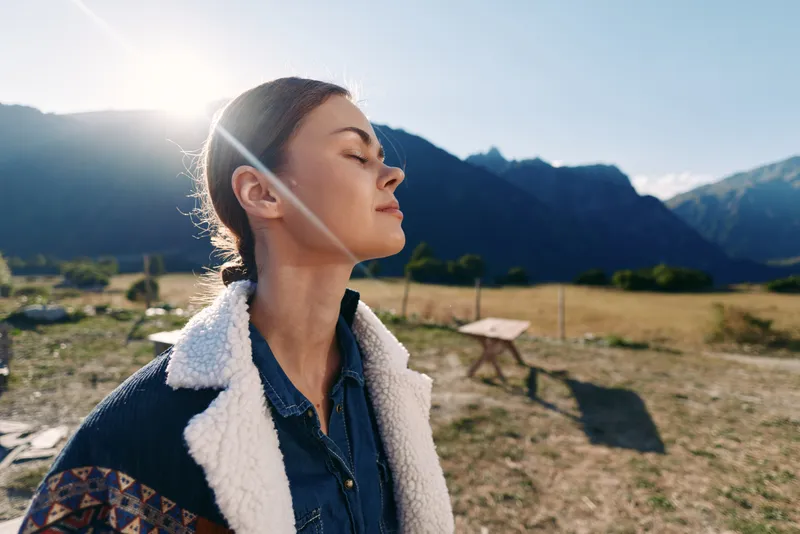 Woman portrait in mountains with closed eyes and sunlight on face, nature outdoors in meadow, serene expression in cozy jacket capturing peaceful travel and fresh air moment.