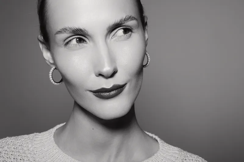 Portrait of a young Caucasian woman with fair skin looking sideways, wearing hoop earrings and a light sweater. Studio lighting highlights facial features and smooth skin.