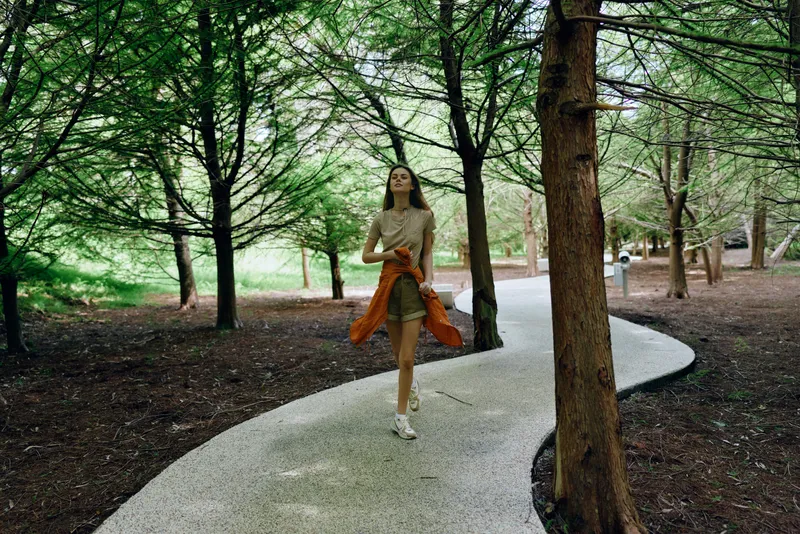 Woman walking on a winding path through tall trees in a park and forest, casual outfit with orange jacket tied at waist, serene nature scene and solitary outdoor stroll.