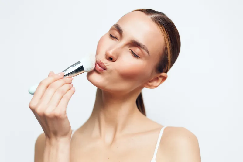 Young woman with clear skin applying makeup with brush, closed eyes, soft expression, natural light, white background, beauty routine, skincare concept