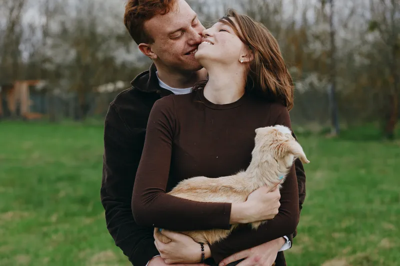 A loving couple embraces a playful goat kid in an open field, capturing intimate connection and joyful companionship in a natural outdoor setting, with soft light creating a serene countryside mood