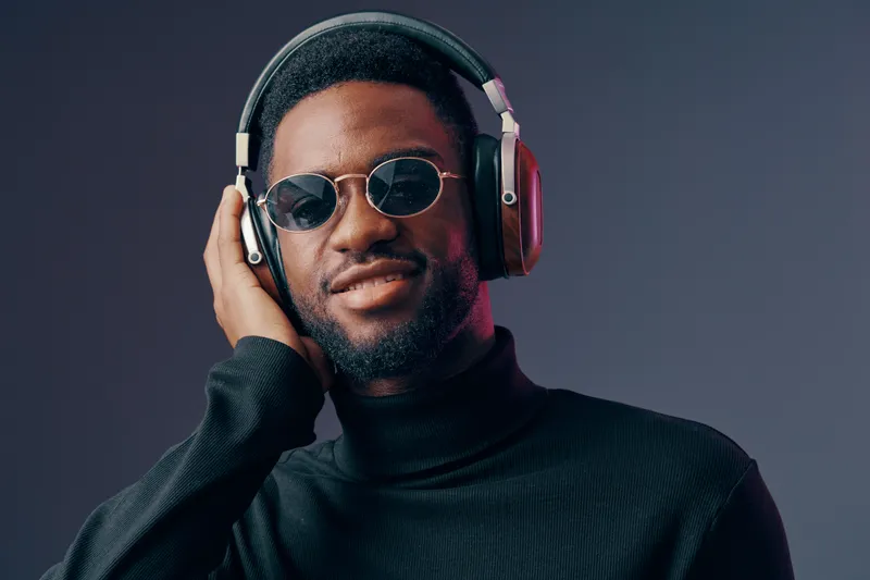 Stylish young Black man smiling while wearing headphones and sunglasses against a dark background, conveying a sense of joy and confidence in music