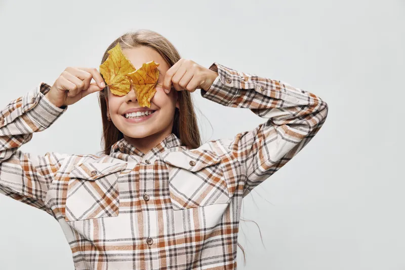 Smiling girl wearing plaid shirt holding yellow autumn leaves in front of her eyes, posing on gray background. Happy child playing with fall foliage, seasonal concept with nature.