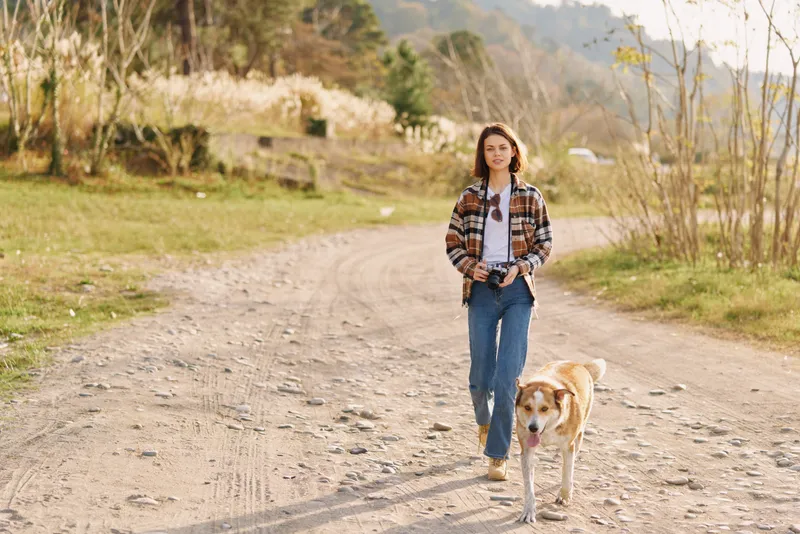 Woman walking on a dirt road with a leash dog in a rural setting, casual attire and plaid shirt, jeans, enjoying nature and daylight with a calm countryside mood.