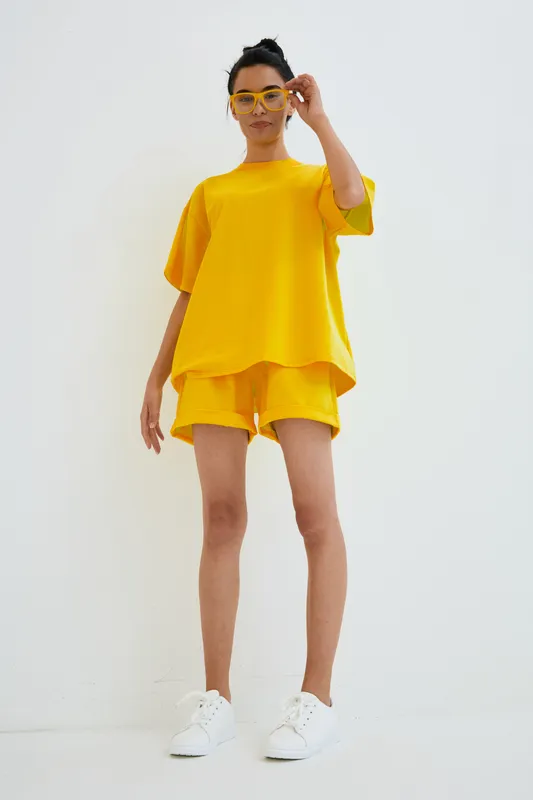 Woman in yellow casual outfit and matching glasses poses confidently against a white background, wearing comfortable shorts and white sneakers, showcasing modern minimal style.