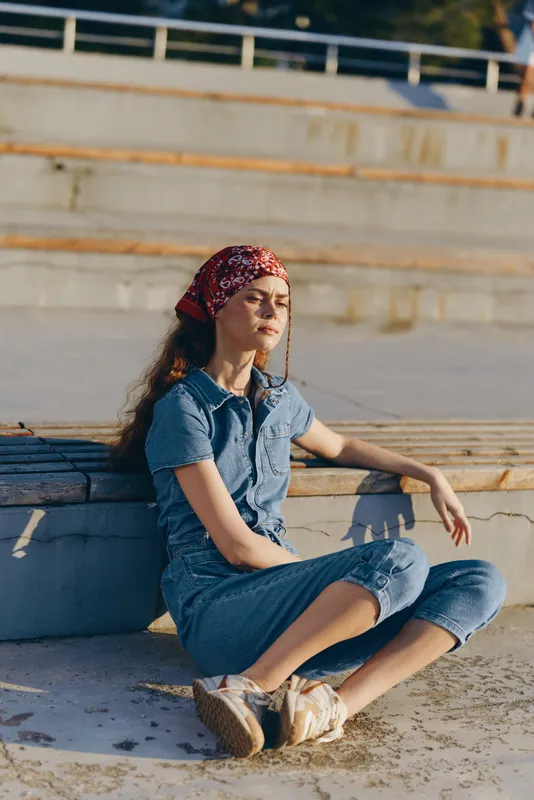 lifestyle woman in modern boho-western denim street style with a red bandana sitting outdoors in warm film color tones, relaxed and thoughtful mood under sunlight