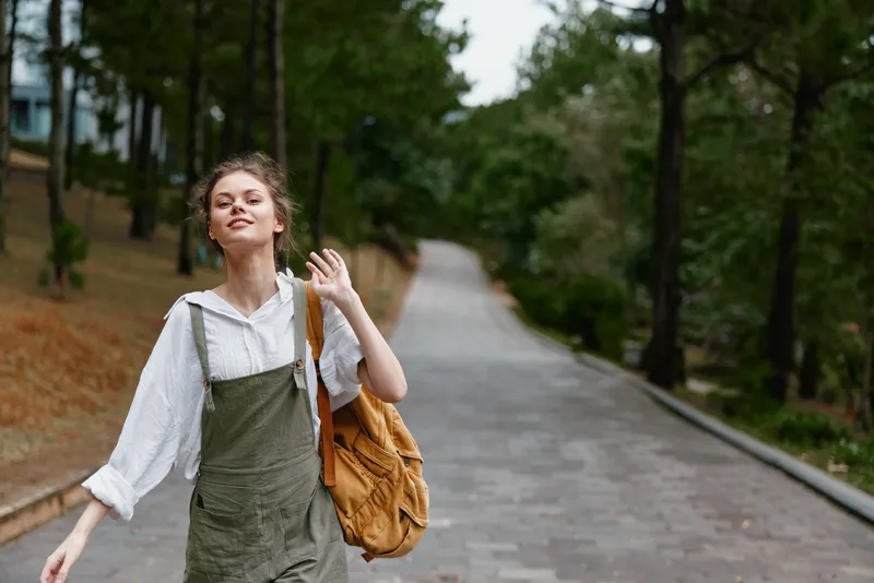 Young woman walking joyfully in a park, wearing a casual outfit with a backpack, showcasing a carefree emotion amidst greenery, ideal for lifestyle concepts