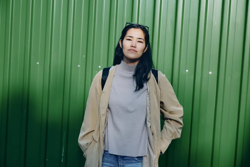 Woman wearing a gray sweater and beige coat stands before a vibrant green wall, showcasing casual urban style, confident pose, and contemporary fashion in natural light.