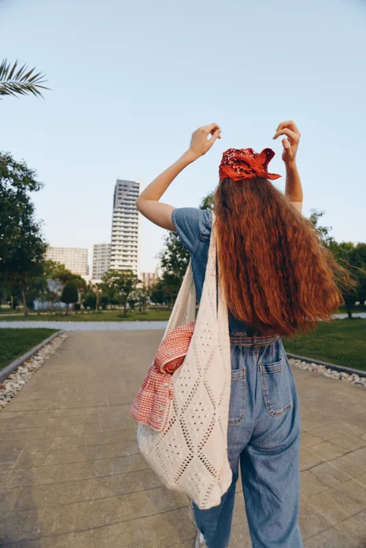 Lifestyle woman with long red hair in modern boho-western denim street style adjusting headscarf outdoors, bright film color, casual urban setting with clear sky.