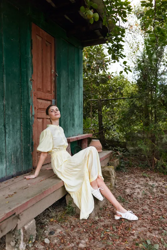Woman in yellow dress sitting on rustic wooden porch of green cabin surrounded by trees relaxed pose natural light authentic inclusive portrait lifestyle countryside calm sunlit vintage evening escape