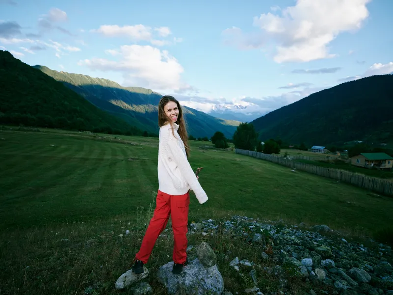 Woman in countryside field wearing whitesweater and redpants smiling near mountains under blue sky outdoors moment showcasing nature meadow rural scenery