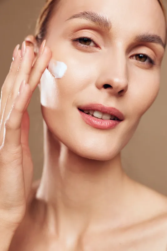 Woman closeup applying facial cream to smooth face skin, beauty skincare routine, gentle touch on cheek, soft studio lighting, calm expression, beige background, personal care