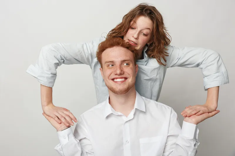 Smiling young man in white shirt with curly red hair looks away while woman with wavy hair leans on his shoulders. Studio portrait on light background. Concept of friendship and happiness.