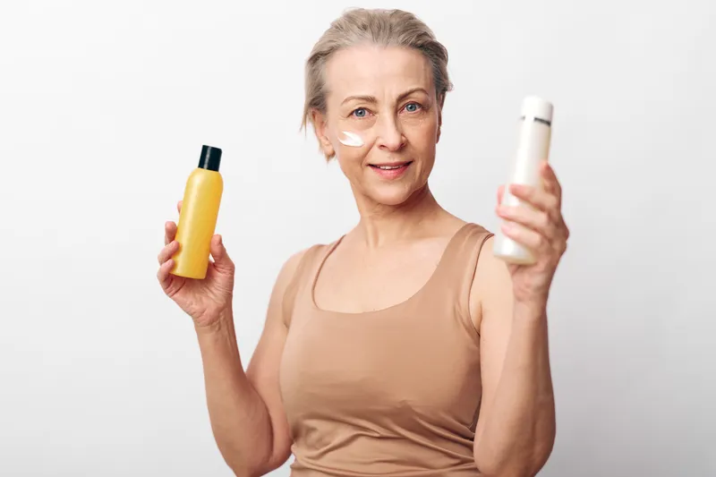 Mature woman with skincare cream on face holds two cosmetic bottles smiling, isolated on white background. Concept of beauty, antiaging, wellness, and natural self-care routine.
