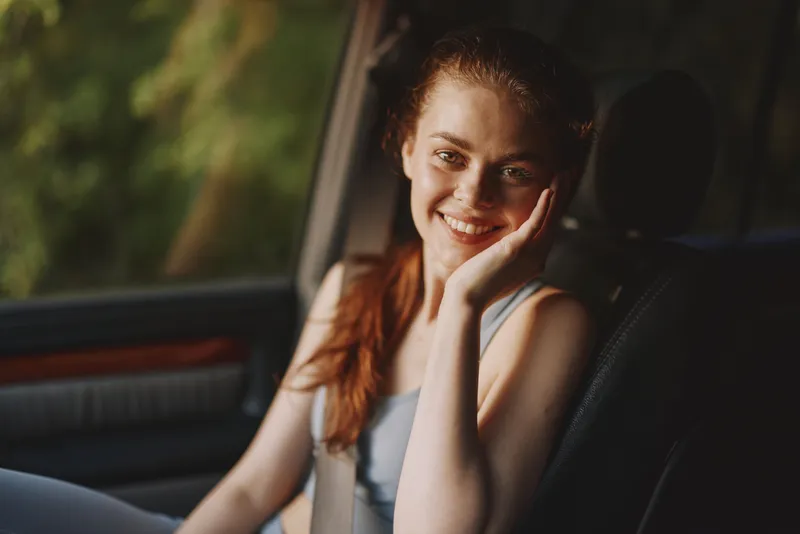 Smiling young woman with red hair sitting in a car, wearing a casual outfit, showcasing a joyful expression against a blurred green background