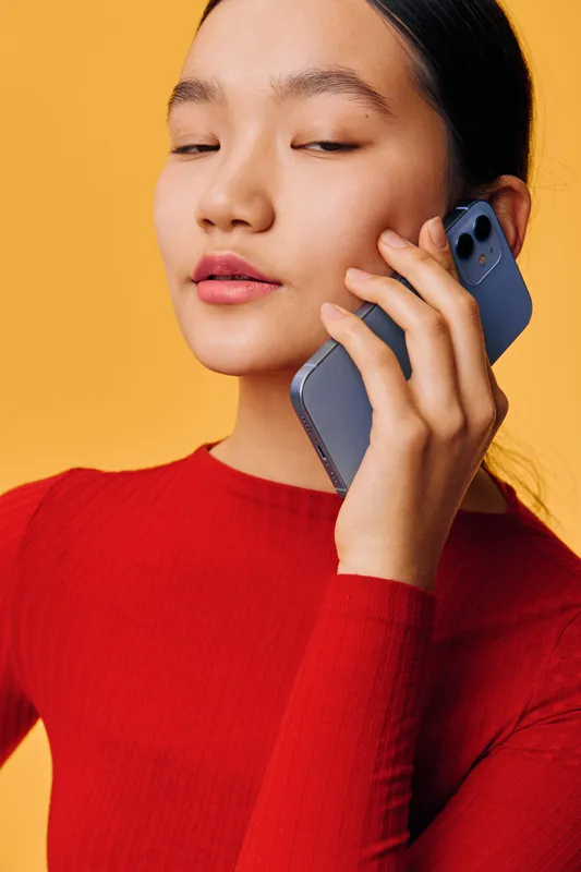 Woman in red sweater talking on smartphone with eyes closed against yellow background. Portrait of young female holding mobile phone for communication and connection.