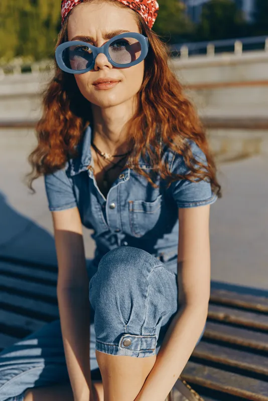 Young woman in blue denim outfit wearing oversized sunglasses sitting outdoors on wooden bench in warm sunlight with confident expression and red curly hair.