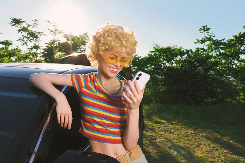 Woman outdoors leans from a car window, wearing a rainbow striped crop top, bright retro color grade, smiles while checking phone, casual lifestyle moment in warm sunlight.