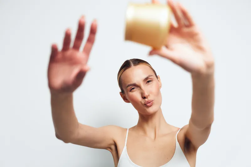 Young woman with clear skin holding skincare cream jar close to camera, smiling with puckered lips. White background, natural makeup, wellness and beauty concept, bright soft lighting.