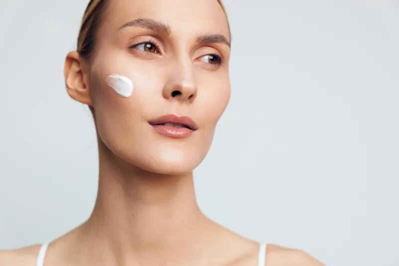 Young woman with fair skin applies white face cream on her cheek while looking thoughtfully to the side against a plain light background.