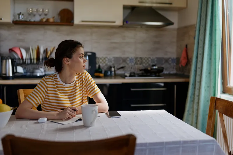Young woman seated at a kitchen table wearing a striped yellow shirt, gazing to the side with a mug in hand. Warm daylight and a cozy home setting convey daily routine and self care.