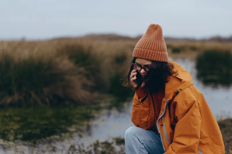 Woman outdoors lifestyle lake beanie jacket autumn sunshine, relaxed pose by shoreline, smiling in soft daylight, warm orange jacket, knit hat and sunglasses, calm countryside background, natural