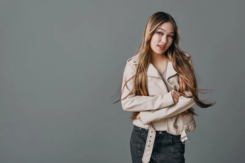 Young woman with long hair wearing light jacket and jeans standing with crossed arms against plain gray background for casual style and confident pose in studio lighting.