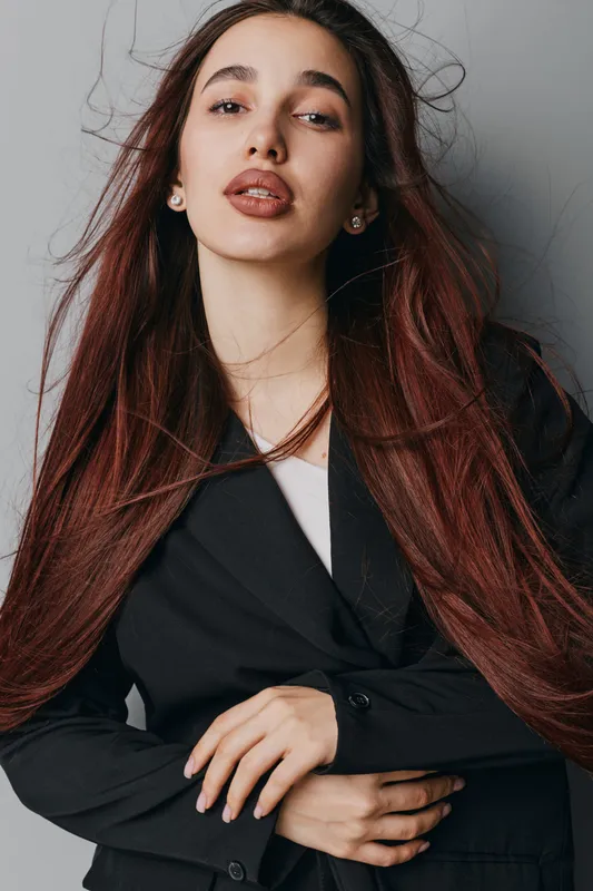 Young woman with long brown hair wearing black blazer posing indoors with confident expression and natural makeup, studio portrait with neutral background and soft lighting.