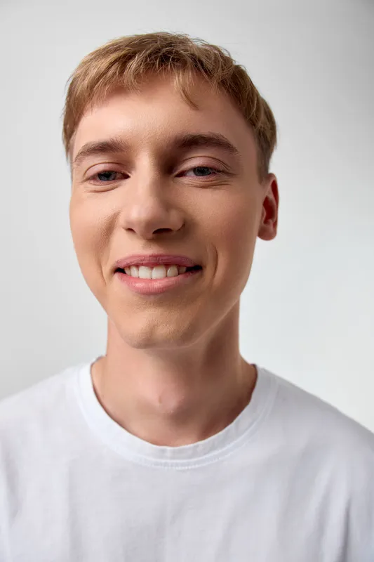 Smiling young man in a white t-shirt against a plain studio backdrop, conveying a friendly, approachable mood and casual everyday style.