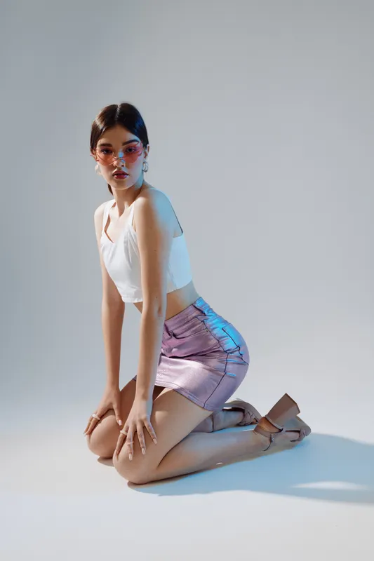Young woman in a white top and shiny pink skirt kneels on the floor wearing stylish sunglasses. Studio shot with soft lighting. Concept of woman beauty fashion and modern style.