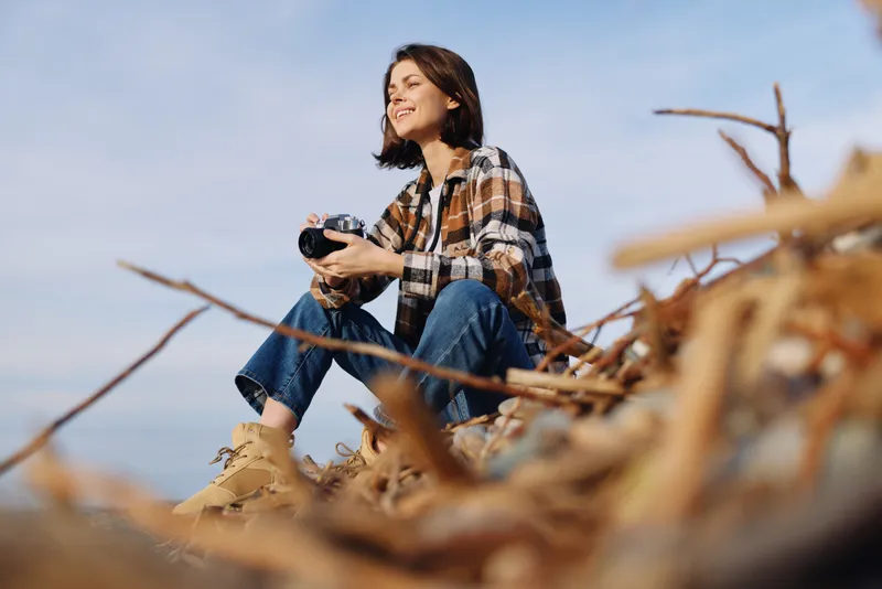 Woman sits outdoors among dried branches, wearing plaid shirt and jeans, holding a camera, enjoying nature and a quiet moment under a clear blue sky.