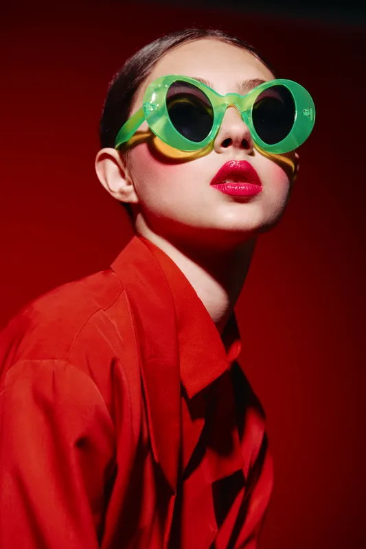 Fashionable woman wearing bright green oversized sunglasses and vivid red lipstick poses confidently against a red background. Concept features bold beauty and creative style.